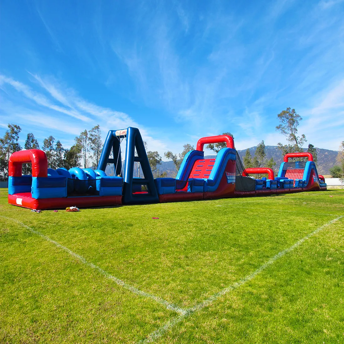 The Rugged Warrior Challenge, a long red and blue inflatable obstacle course, set up on a grass field under a bright blue sky